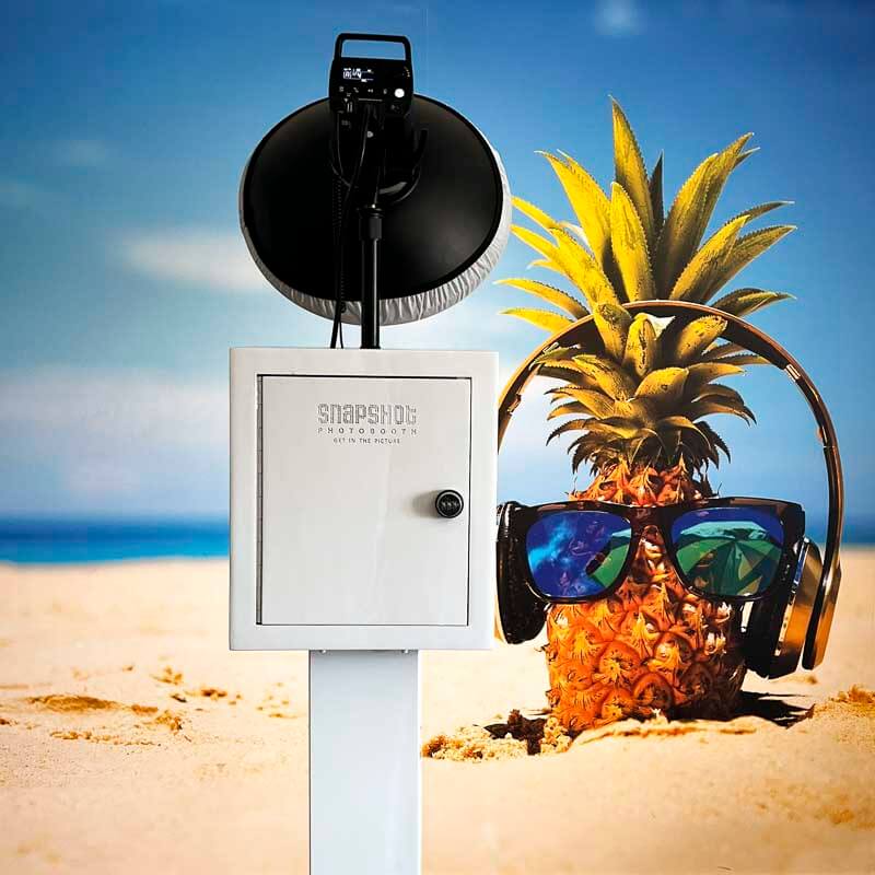 Snapshot-Photobooth-Malta-Jr-About-Us A photo booth pointed at a pineapple wearing head phones and sunglasses on a beach