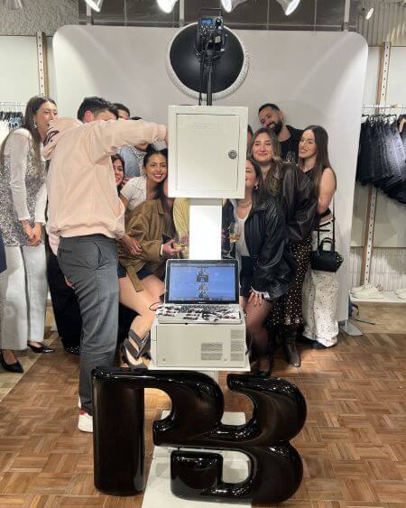 Pull-and-bear-malta-30th-anniversary-photo-booth-group Group of people gathered around a photo booth machine at a retail event, posing for a picture in a clothing store.