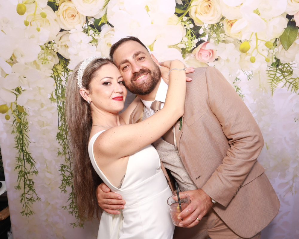 Just Married Elysia & Stefan-01 Bride Elysia and groom Stefan smiling and hugging in front of a white and pink floral wedding backdrop.