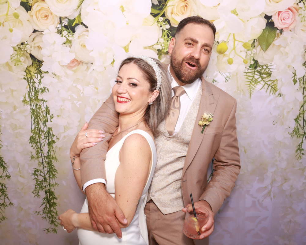 Just Married Elysia & Stefan-02 Bride Elysia and groom Stefan smiling and posing together with wedding guests in front of a white and pink floral backdrop.