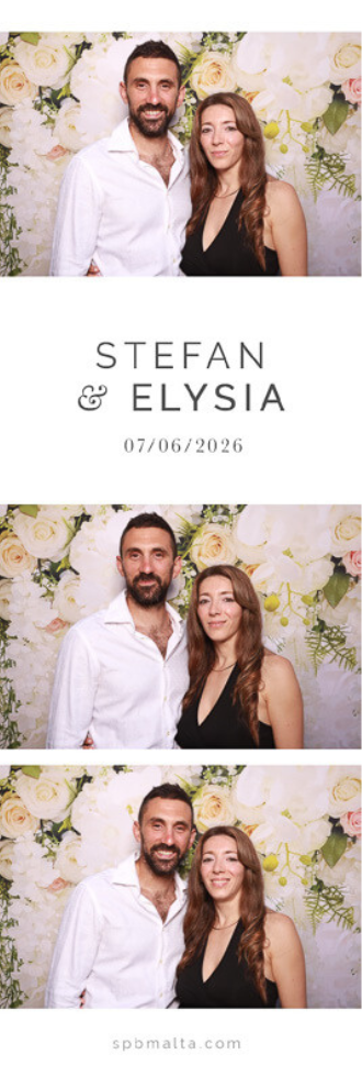 Stefan & Elysia-01 A man in a white shirt and a woman in a black dress posing together in front of a white and pink floral wedding backdrop.