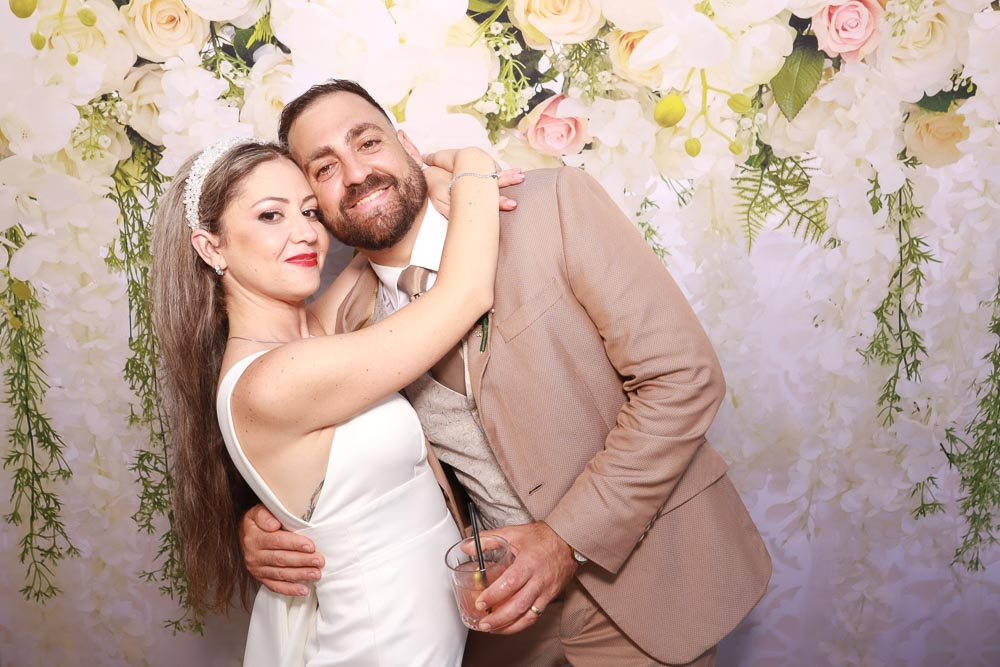 wedding-photo-booth-malta-bride-and-groom-posing Bride Elysia and groom Stefan smiling and hugging in front of a white and pink floral wedding backdrop.