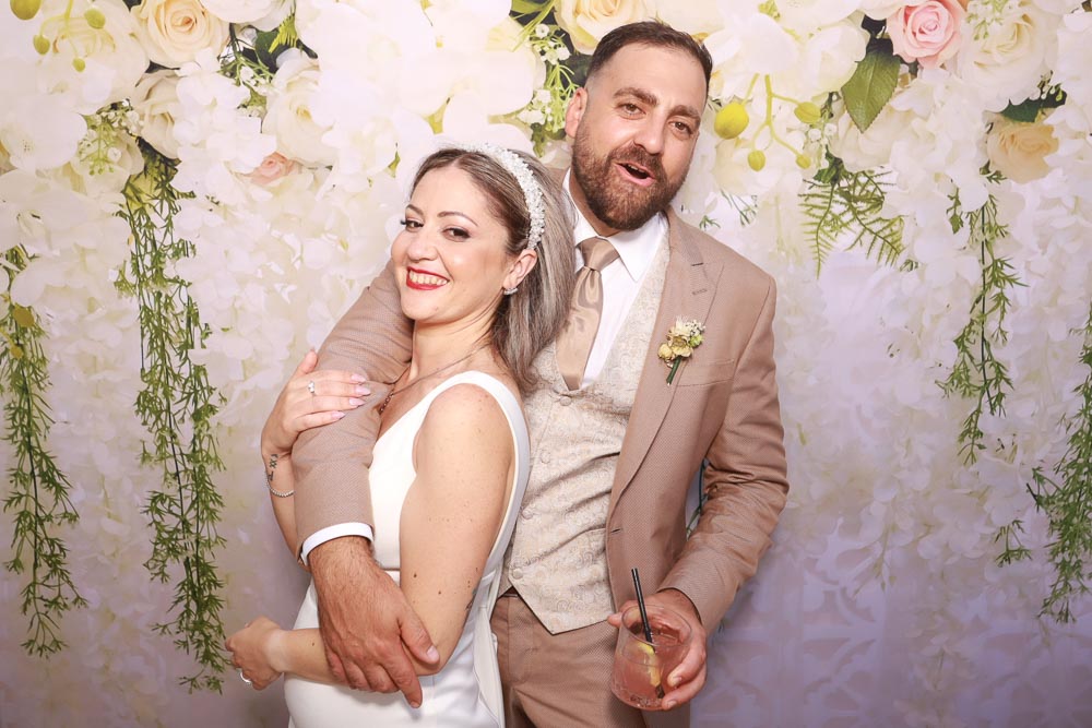 wedding-photo-booth-malta-bride-and-groom-smiling-together. Bride Elysia and groom Stefan smiling and posing together with wedding guests in front of a white and pink floral backdrop.