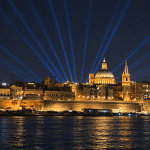 Valletta waterfront at night illuminated by golden lights and blue searchlights across the sky.