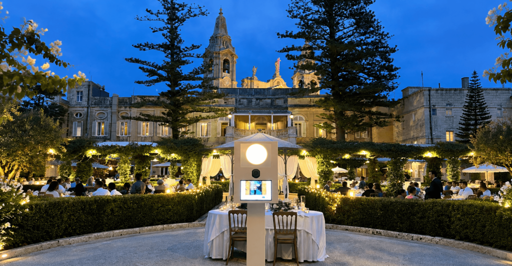Photo Booth at a Wedding in Malta White photo booth in front of a candlelit garden wedding dinner at a historic Maltese palazzo, with bell towers and pine trees at dusk.