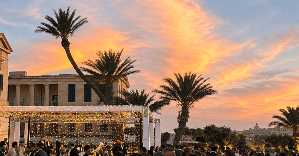 Sunset Wedding Reception in Malta Wedding guests gather by a fairy-lit stage at sunset in Malta, with palm trees, limestone buildings and an orange sky.